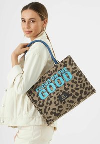 Tote-Bag mit Leopardenmuster, blauer "FOCUS ON THE GOOD" Schriftzug, blauen Griffen und einem schwarzen Etikett mit einem Logo. Strukturierter Stoff.