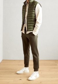 Man som bär en olivgrön vadderad väst, beige knapp-up skjorta, matchande olivgröna byxor, vita strumpor och vita sneakers står på ett trägolv.