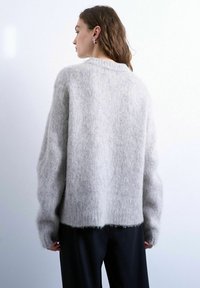 Femme aux longs cheveux bruns portant des boucles d'oreilles argentées en forme d'anneau et un pull ample et doux de couleur gris clair, vue de dos sur un fond uni.