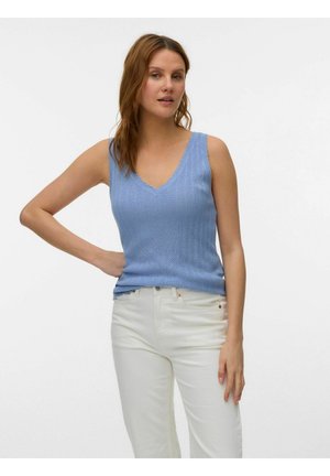 Femme aux cheveux châtains clairs portant un haut bleu sans manches en maille à col en V et un pantalon blanc, debout avec une main sur la hanche.