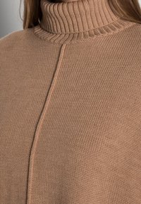 Sweter z golfem dzianinowym w jasnobrązowym kolorze. Posiada prążkowany kołnierz oraz gładką fakturę z bezszwowym wykończeniem i centralnym detalem w postaci ściegu.