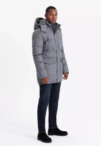 Giubbotto puffo grigio con cappuccio, dotato di chiusura con cerniera e tasche frontali. Indossato sopra jeans in denim scuro e stivali neri.