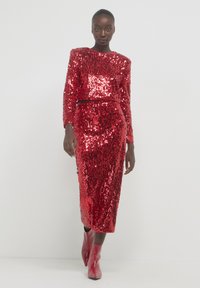 Completo a due pezzi rosso con paillettes, maniche lunghe e silhouette aderente. La gonna arriva a metà polpaccio, abbinata a stivali alla caviglia rossi.