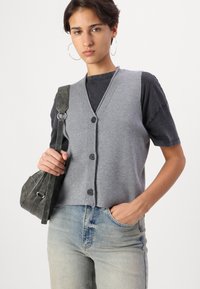 Cardigan senza maniche grigio con scollatura a V e quattro bottoni neri, indossato sopra una maglietta nera, abbinato a jeans azzurri chiari e a una borsa piccola.