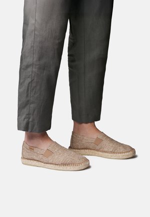 Person trägt beige gewebte Espadrilles ohne Schnürung und dunkelgraue, gekürzte Hosen, steht vor hellem Hintergrund.