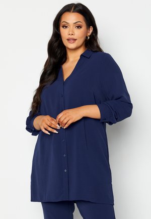 Chemise bleu marine à boutons avec une coupe décontractée, décolleté en V, manches longues retroussées jusqu'aux coudes et tissu doux et texturé.