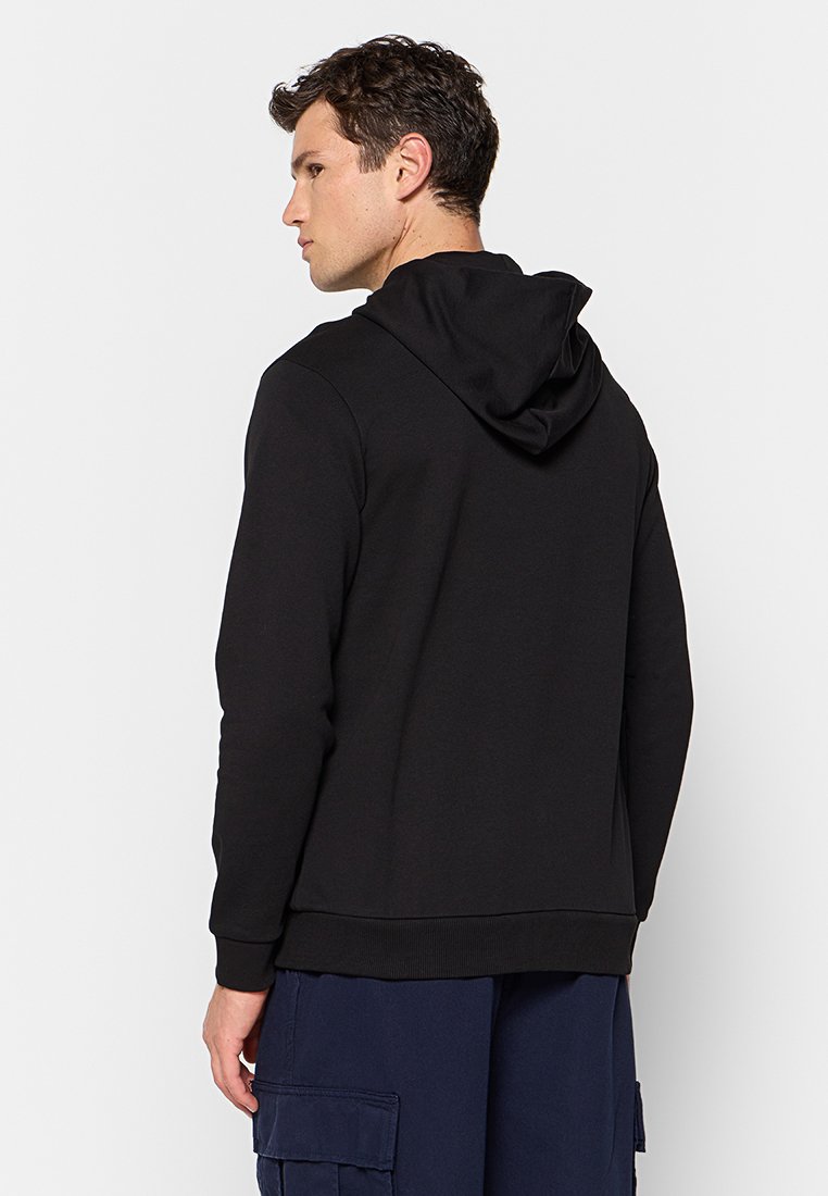 Homme de profil portant un sweat à capuche noir et un pantalon cargo bleu foncé devant un fond clair.