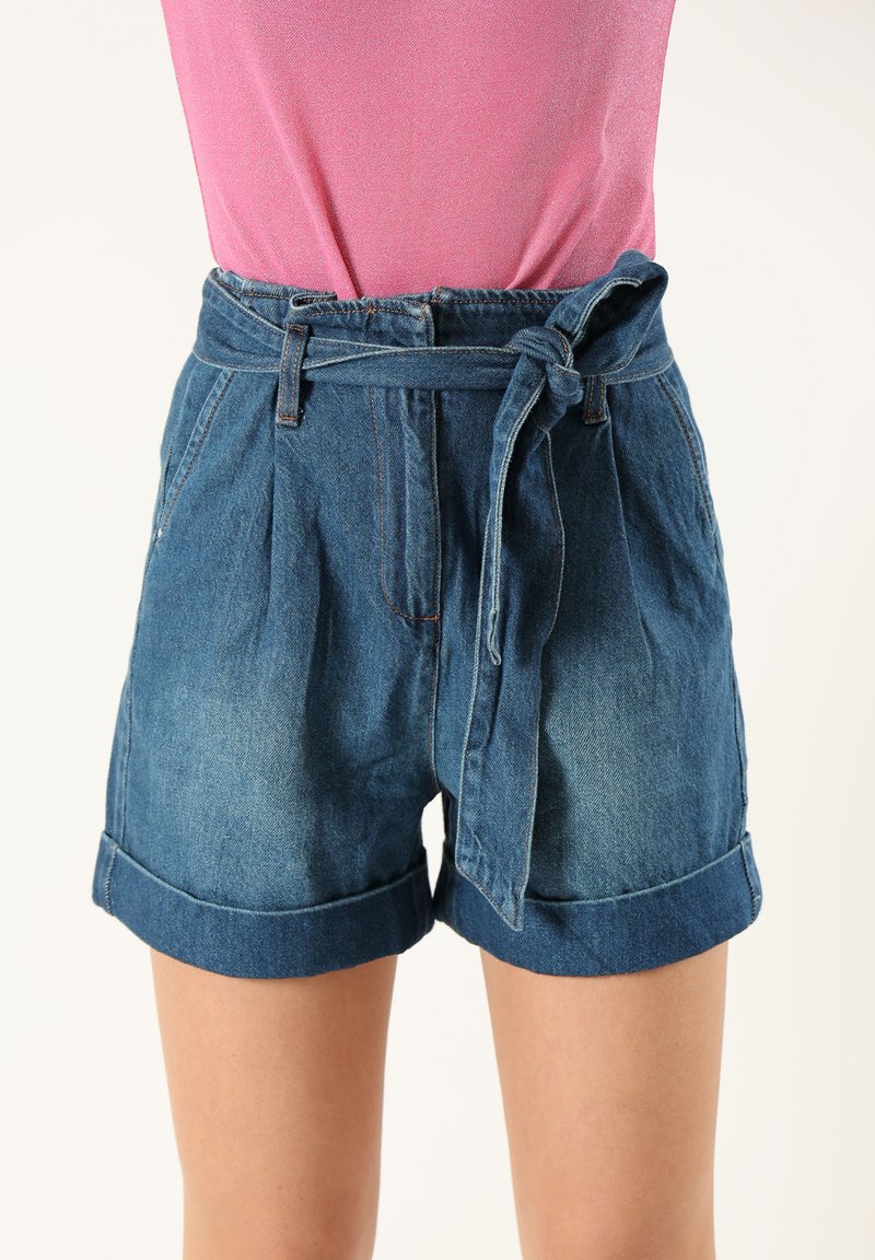 Shorts de mezclilla de cintura alta, con un cinturón anudado, frente con pliegues y dobladillos enrollados. La tela es de un azul medio con un ligero deslavado.