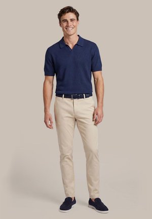 Glunderende man staand, gekleed in een marineblauw poloshirt met korte mouwen, beige chino’s met riem en marineblauwe instapschoenen tegen een effen achtergrond.