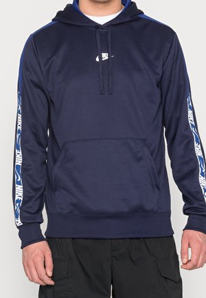 Mężczyzna w granatowej bluzie Nike z białym logo na piersi i markowymi niebiesko-białymi paskami na rękawach, w połączeniu z czarnymi bojówkami.