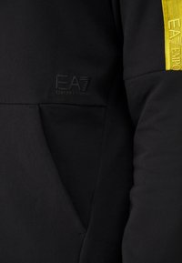 Černý rukáv mikiny s přední kapsou, na kterém se nachází černé embosované logo "EA7 Emporio Armani" a žlutý pruh s částečným textem "EA7 Emporio Armani".