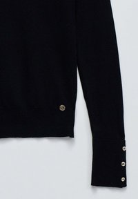 Pull en tricot noir avec un détail de logo rond à l'ourlet et des accouts en bouton sur les poignets. Présente un design ajusté et une texture côtelée.