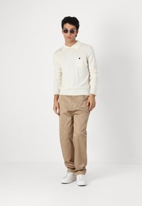 Polo de manga longa em creme claro com um bolso no peito, combinado com umas calças bege e ténis brancos, usado por uma pessoa com óculos de sol.