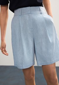 Shorts plissettati azzurri in tessuto strutturato, con tasche laterali e una cintura liscia, mostrati dalla vita in giù.