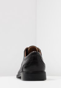 Chaussure en cuir noir avec une texture lisse, un design arrondi et des coutures d'accent sur le côté. La semelle est texturée et légèrement surélevée.