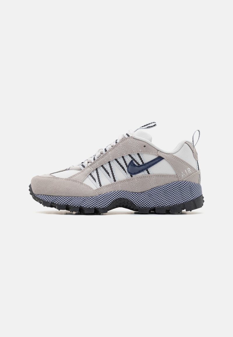 Baskets basses blanches et grises avec des rayures noires, un swoosh bleu marine, une semelle intermédiaire texturée et une semelle extérieure noire robuste.