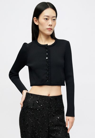 Femme jeune en cardigan noir court à boutons et pantalon paillettes noirs taille haute, se tenant avec une main sur la hanche devant un fond blanc.