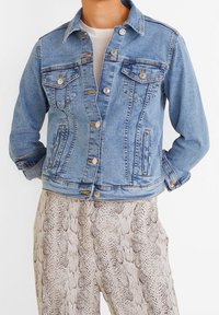 Veste en denim bleu clair avec un col classique, deux poches poitrine, boutons en métal argenté et une silhouette légèrement ajustée. Portée par-dessus un pantalon en cuir de serpent à motifs.