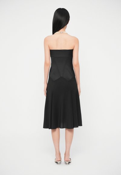 Žena s dlhými čiernymi vlasmi, ktorá má na sebe čierne midi šaty bez ramienok, perlový náhrdelník a biele vysoké podpätky, stojaca na bielom pozadí.