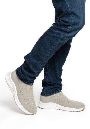 Person trägt beige Strick-Slipper-Sneaker und dunkelblaue Jeans, steht mit einem leicht angehobenen Fuß.