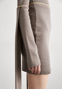 Gros plan sur un bras et un torse portant une robe en maille côtelée taupe avec des manches longues dépassant le poignet et une fente latérale.