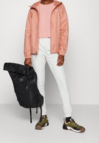 Chaqueta rosa ligera, pantalones deportivos blancos y mochila negra. Cuenta con puños elásticos y capucha, junto con zapatillas texturizadas y transpirables.