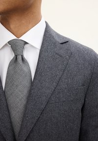 Giacca da abito grigia in lana con rever a punta, abbinata a una camicia bianca e a una cravatta grigia strutturata. Il tessuto appare morbido e su misura.