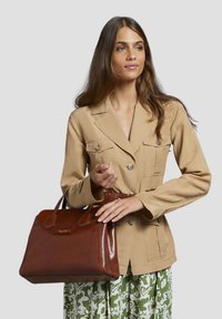 Borsa a mano in pelle marrone con finitura lucida, manici corti, forma rettangolare e branding sottile. Il modello indossa una giacca beige e una gonna a fantasia verde.