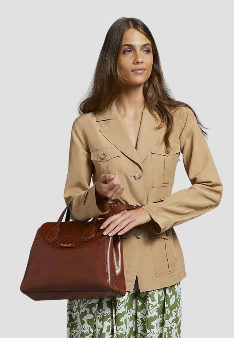 Borsa a mano in pelle marrone con finitura lucida, manici corti, forma rettangolare e branding sottile. Il modello indossa una giacca beige e una gonna a fantasia verde.