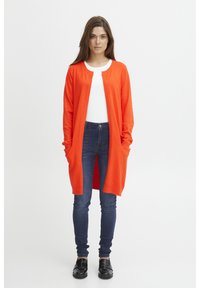 Long cardigan ouvert en tricot orange vif avec des détails côtelés, associé à un haut blanc, un jean noir et des chaussures noires.