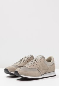 Beige semišové tenisky s síťovinovými prvky, kulatou špičkou, šněrovacím designem, polstrovaným límcem a černou gumovou podešví. Obsahují velké logo 'N'.