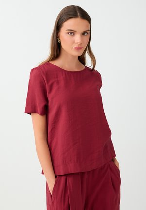 Jeune femme aux longs cheveux bruns portant un haut bordeaux à manches courtes et un pantalon assorti, debout les mains dans les poches, sur fond blanc.