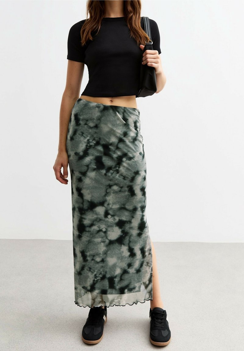 Femme debout à l'intérieur portant un crop top noir, une jupe longue verte tie and dye avec fente sur le côté, des baskets noires et un sac à bandoulière noir.