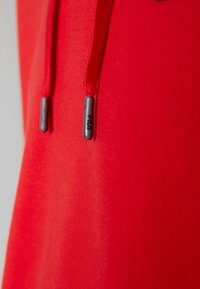 Primer plano de una tela roja con dos cuerdas rojas, cada una rematada con ojales grises que presentan un pequeño logo negro.