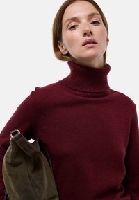 Pull col roulé bordeaux en tissu tricoté doux, avec un col haut côtelé. Un sac vert avec une sangle marron est tenu sur le côté.