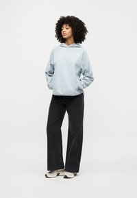 Ανοιχτό μπλε φούτερ με κουκούλα και μπροστινή τσέπη με λογότυπο, συνδυασμένο με μαύρο παντελόνι με φαρδιά πόδια και μπεζ αθλητικά παπούτσια. Απλό φόντο.