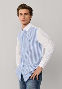 Jeune homme aux cheveux bruns portant une chemise blanche à rayures verticales bleues sur le devant et un jean bleu, posant avec une main sur la hanche.