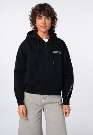 Frau mit lockigem Haar, die einen schwarzen Hoodie mit Reißverschluss trägt, mit Text auf Brust und Ärmel, kombiniert mit hellgrauen Hosen, vor weißem Hintergrund.