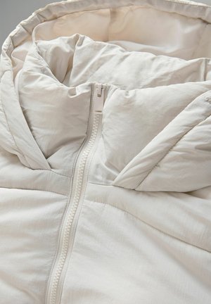 Ljus beige pufferjacka med hög krage, med en central dragkedja och mjukt, strukturerat tyg. Kvadret design med synliga sömmar.