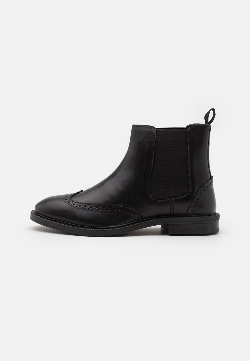 Bottine Chelsea en cuir noir avec panneaux latéraux élastiques, détails brogue, talon bas et languette de tirage sur fond gris clair.