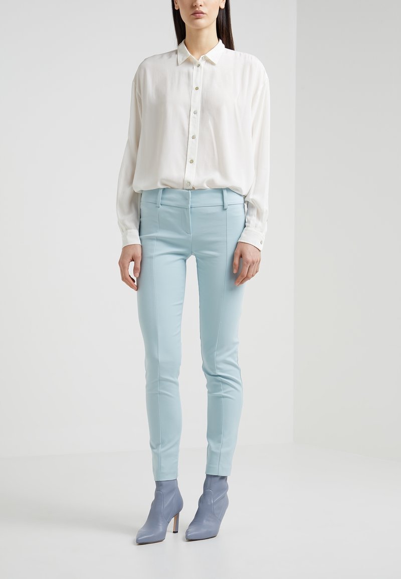 Une chemise blanche à manches longues avec des boutons est associée à un pantalon slim bleu clair et des bottines grises, présentant un design élégant et une texture lisse.