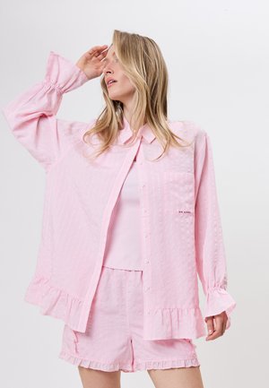 Žena nosící světle růžovou texturovanou košili s dlouhým rukávem na knoflíky s volánkovými lemy a odpovídající šortky, pózuje s rukou u čela.