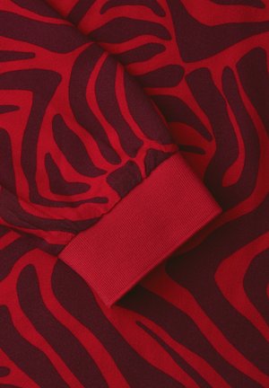 Tissu rouge avec des rayures abstraites semblables à celles d'un zèbre dans une teinte bordeaux foncé, accompagné d'un détail de couture côtelée au niveau du poignet d'une manche de vêtement.