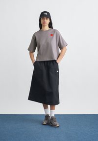 Νέα γυναίκα που στέκεται με τα χέρια στις τσέπες, φορά γκρι κοντοκομμένο μπλουζάκι, μαύρη μίντι φούστα, λευκές κάλτσες, γκρι αθλητικά παπούτσια και μαύρο καπέλο.