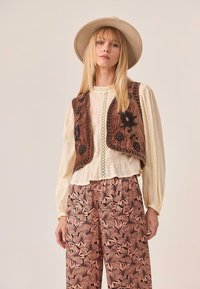 Mujer con sombrero de ala ancha beige, blusa de manga larga crema con detalles de encaje, chaleco bordado marrón y pantalones de pierna ancha rosas con flores.