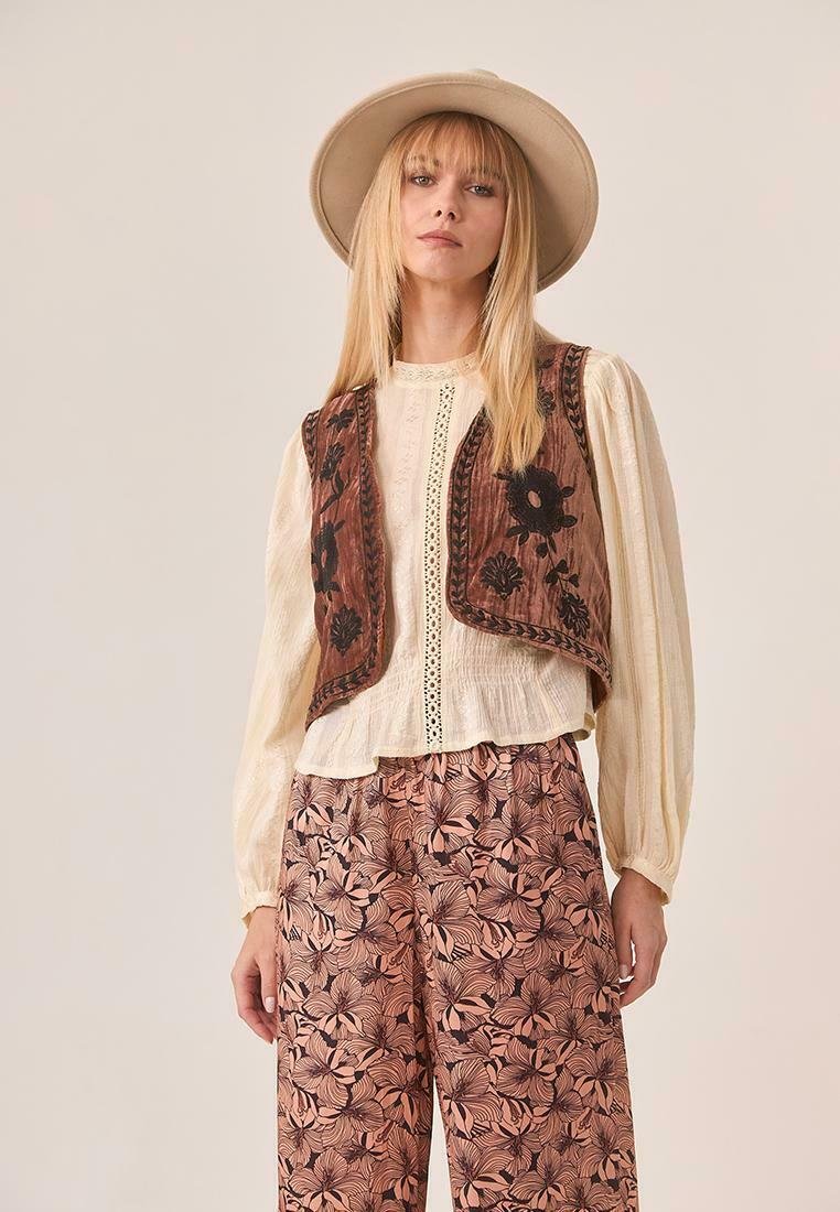 Mujer con sombrero de ala ancha beige, blusa de manga larga crema con detalles de encaje, chaleco bordado marrón y pantalones de pierna ancha rosas con flores.