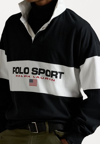 Zwart-wit rugbyshirt met een opgestikte kraag. Bevat de tekst "POLO SPORT RALPH LAUREN" en een Amerikaanse vlag in het rood. Gemaakt van katoenmix.