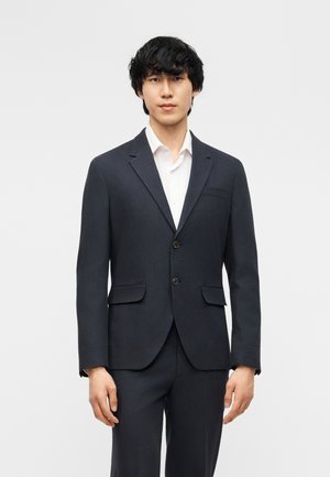 Ung mann med svart krøllete hår iført en mørk dressjakke med to knapper over en hvit skjorte, stående foran en ensfarget lys bakgrunn.