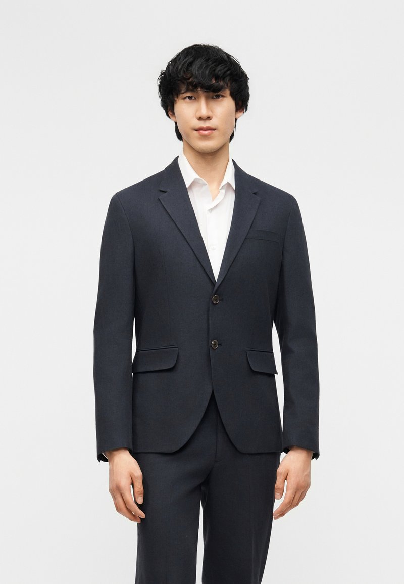 Νεαρός άνδρας με μαύρα σγουρά μαλλιά, ντυμένος με σκούρο σακάκι με δύο κουμπιά πάνω από ένα λευκό πουκάμισο, στέκεται μπροστά από ένα απλό φωτεινό φόντο.