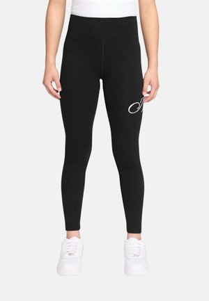 Mujer con leggings negros de tiro alto con logo en letras blancas en la parte superior del muslo, combinados con zapatillas blancas y una camiseta blanca.